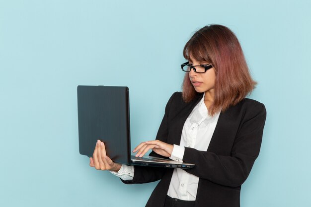 Weibliche Büroangestellte der Vorderansicht im strengen Anzug unter Verwendung des Laptops auf der hellblauen Oberfläche