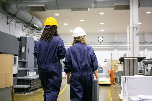 Weibliche Fabrikarbeiter in Schutzhelmen und Overalls, die auf dem Werksgelände gehen und sprechen und Werkzeugkasten tragen