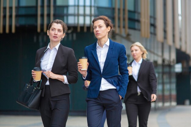 Weibliche Fachleute mit Kaffeetassen aus Papier, die Büroanzüge tragen, zusammen in der Stadt spazieren gehen, reden, über ein Projekt diskutieren oder sich unterhalten. Vorderansicht. Geschäftsfrauen im Freien Konzept