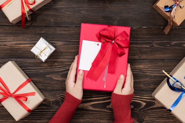 Weibliche Hände, die eine kleine Schachtel mit einem Geschenk unter festlichen Winterdekorationen auf einer weißen Tischplatte halten. Flache Zusammensetzung für Geburtstag, Weihnachten oder Hochzeit.