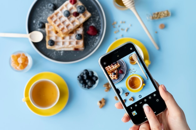 Weibliche Hände, die Foto von Waffeln mit Früchten und einer Tasse Tee machen