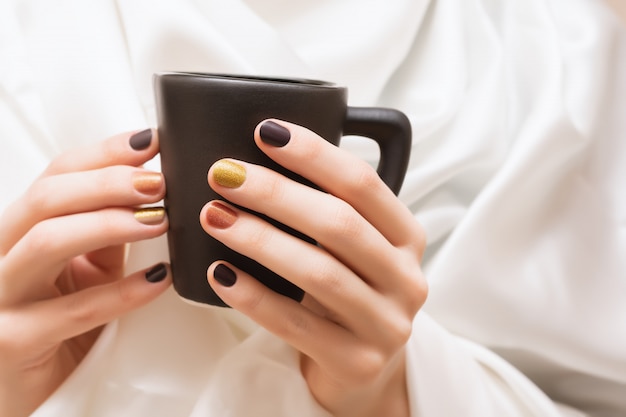 Weibliche Hände mit braunem Nageldesign, das schwarze Tasse hält.