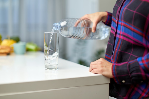 Weibliche hand, die trinkwasserflasche hält und wasser in glas auf dem tisch auf küchenhintergrund gießt.