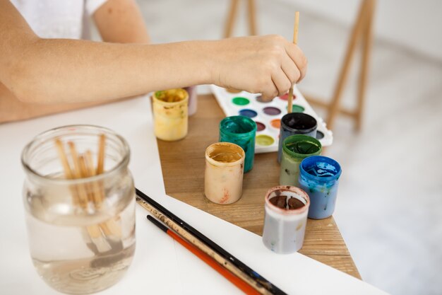 Weibliche Hand hält Pinsel und vertieft ihn in Farbe. Kindermalerei unter Verwendung von Aquarell während des Unterrichts im Kunstraum.