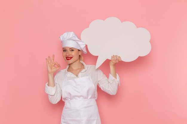 Weibliche Konditorin der Vorderansicht in der weißen Abnutzung, die ein großes weißes Zeichen hält, das auf dem rosa Wandkochjobküchenküchenlebensmittel lächelt