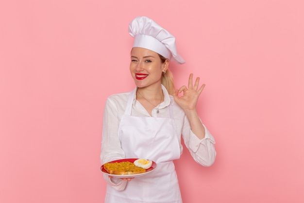 Weibliche Konditorin der Vorderansicht in der weißen Verschleißhalteplatte mit dem Essen auf dem rosa Schreibtischkochjobküchenküchenlebensmittel