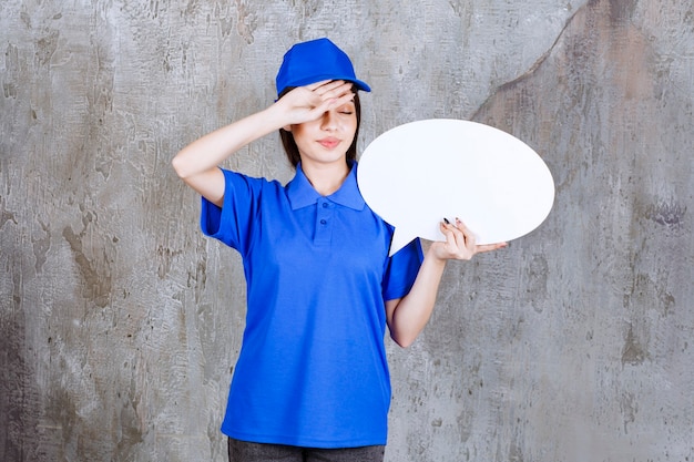 Weibliche Servicemitarbeiterin in blauer Uniform, die eine ovale Infotafel hält und müde aussieht.