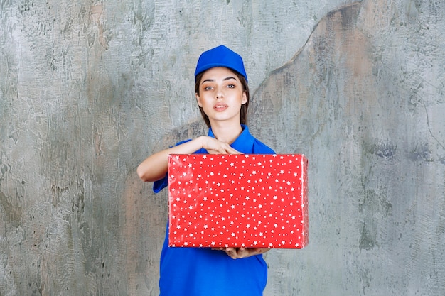 Weibliche servicemitarbeiterin in blauer uniform, die eine rote geschenkbox mit weißen punkten darauf hält.