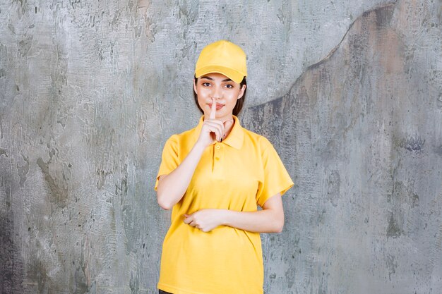 Weibliche Servicemitarbeiterin in gelber Uniform, die auf Betonmauer steht und um Stille bittet.