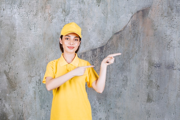 Kostenloses Foto weibliche servicemitarbeiterin in gelber uniform, die auf betonwand steht und auf die rechte seite zeigt.