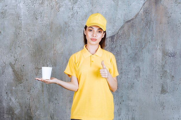 Weibliche Servicemitarbeiterin in gelber Uniform, die einen Plastikbecher hält und ein positives Handzeichen zeigt.