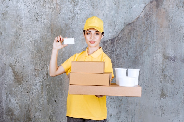 Weibliche Servicemitarbeiterin in gelber Uniform, die einen Vorrat an Pappkartons und Plastikbechern zum Mitnehmen hält, während sie ihre Visitenkarte vorlegt