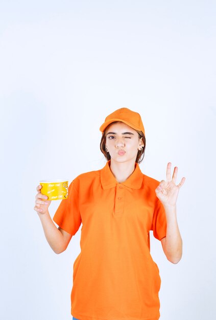 Weibliche Servicemitarbeiterin in orangefarbener Uniform, die eine gelbe Tasse zum Mitnehmen hält und den Geschmack genießt