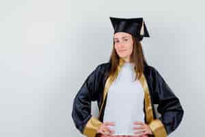 Kostenloses Foto weiblicher absolvent, der hände auf taille in akademischer kleidung hält und stolz aussieht. vorderansicht.