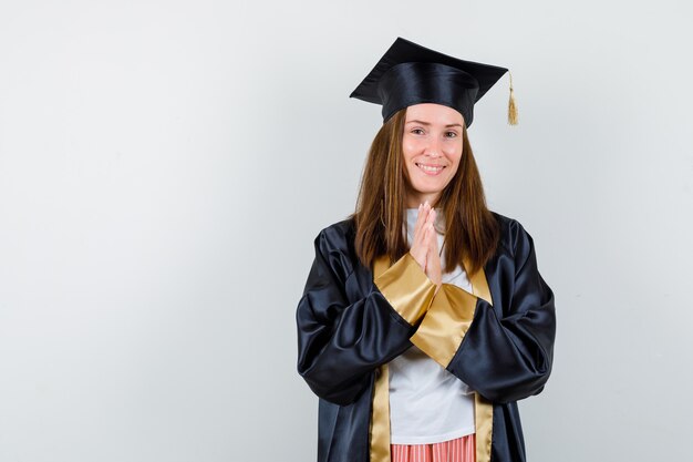 Weiblicher Absolvent in der akademischen Kleidung, die Hände in der Gebetsgeste hält und lustig, Vorderansicht schaut.