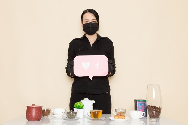 weiblicher Barista in der schwarzen Hemdhose mit kaffeebraun getrockneten Tee-Ausrüstungsbestandteilen in der schwarzen sterilen Maske an der weißen Wand