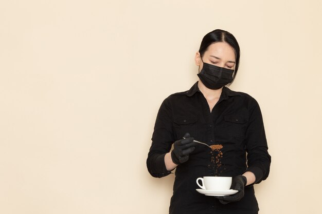weiblicher Barista in der schwarzen Hemdhose mit kaffeebraun getrockneten Tee-Ausrüstungsbestandteilen in der schwarzen sterilen Maske an der weißen Wand