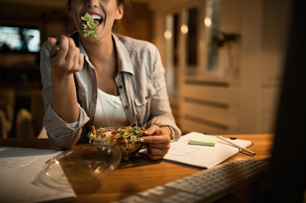 Weiblicher Freiberufler, der Computer benutzt und nachts zu Hause Salat isst