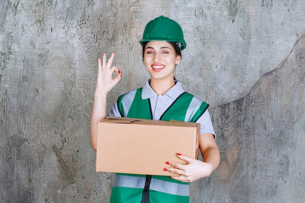 Weiblicher Ingenieur im grünen Helm, der einen Karton hält und Zufriedenheitszeichen zeigt.