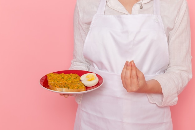 Weiblicher Konditor der Vorderansicht in der weißen Trage-Halteplatte mit Toast und Ei auf der rosa Wandküchenküchennahrung