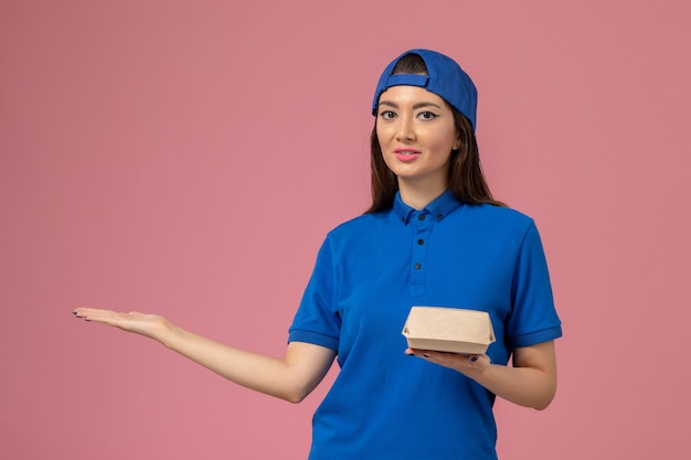 Weiblicher Kurier der Vorderansicht im blauen Uniformumhang, der kleines Lieferpaket auf rosa Wand hält, Mitarbeiterlieferarbeitsarbeitsarbeitermädchenfirma