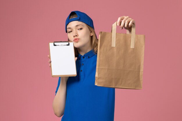 Weiblicher Kurier der Vorderansicht im blauen Uniformumhang, der Lieferpaket und Notizblock denkt, der auf rosa Wand, Mitarbeiterdienstzustellung denkt