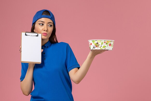 Weiblicher Kurier der Vorderansicht im blauen Uniformumhang, der Notizblock mit Lieferschüssel auf hellrosa Wand hält, Servicemitarbeiter-Lieferarbeit