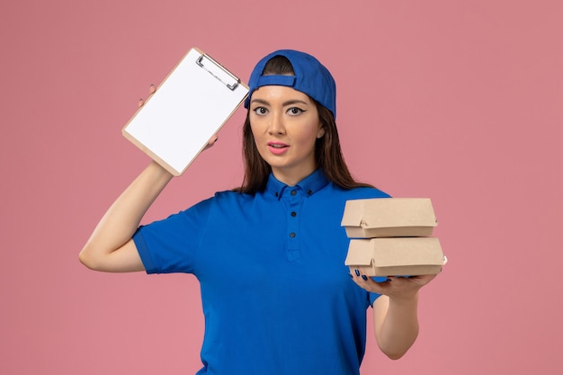 Weiblicher Kurier der Vorderansicht im blauen Uniformumhang, der Notizblock und kleine Lieferpakete auf hellrosa Wand hält, Servicemitarbeiter-Lieferarbeit