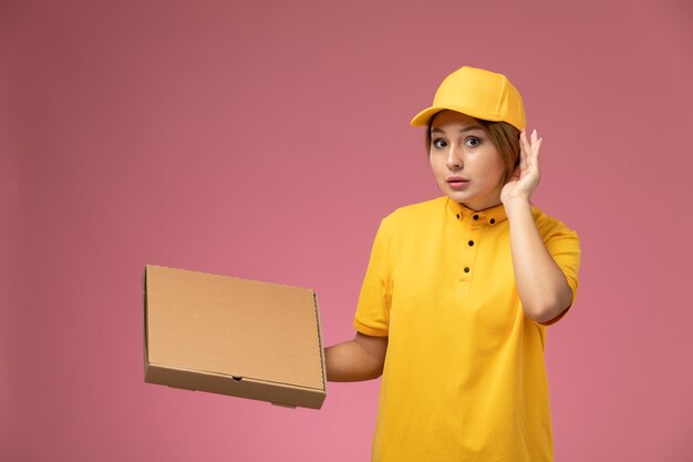 Weiblicher Kurier der Vorderansicht im gelben einheitlichen gelben Umhang, der Lebensmittelpaket hält, das versucht, auf der rosa Schreibtischuniform-Lieferarbeitsfarbe zu hören