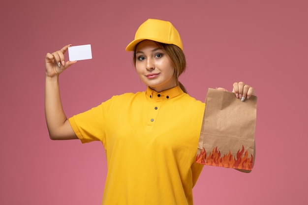 Weiblicher Kurier der Vorderansicht im gelben einheitlichen gelben Umhang, der Lebensmittelpaket mit weißer Karte auf dem rosa Hintergrunduniformlieferarbeitsfarbjob hält