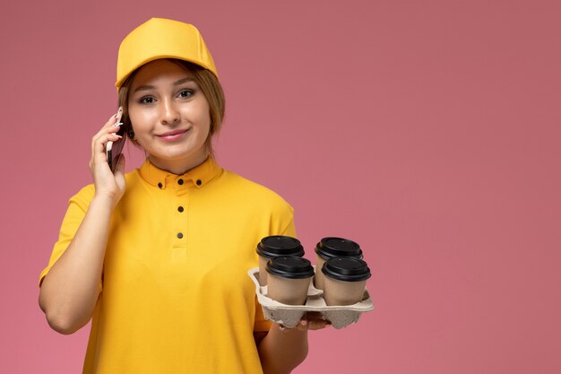 Weiblicher Kurier der Vorderansicht im gelben Uniformgelbumhang, der Plastikkaffeetassen hält, die am Telefon auf der rosa Hintergrunduniform-Lieferfarbe sprechen
