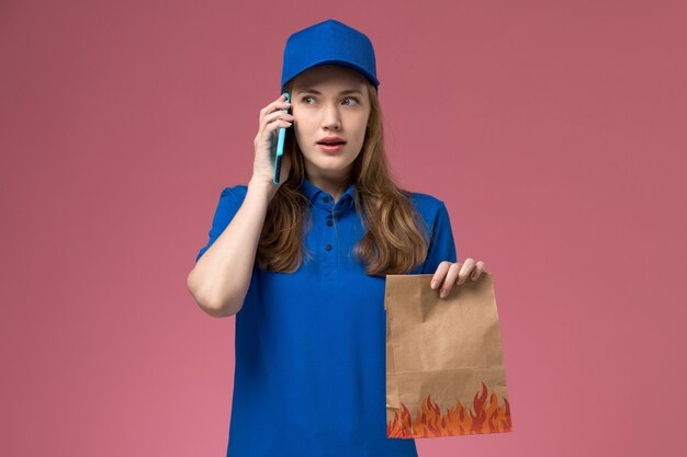 Weiblicher Kurier der Vorderansicht in der blauen Uniform, die am Telefon spricht, während Lebensmittelpaket auf rosa hellem Schreibtischdienstuniformfirmenjob hält
