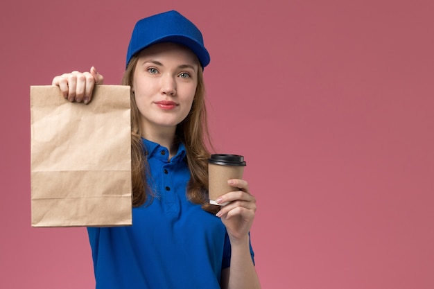 Weiblicher kurier der vorderansicht in der blauen uniform, die braune kaffeetasse mit lebensmittelpaket und lächeln auf hellrosa schreibtischdienstuniform hält, die firma liefert