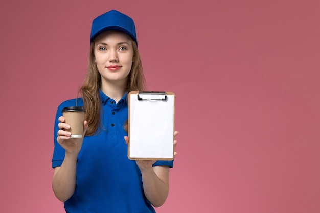 Weiblicher Kurier der Vorderansicht in der blauen Uniform, die braune Kaffeetasse mit Notizblock auf der hellrosa Boden-Dienstjobuniform-Lieferfirma hält