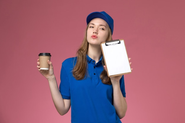 Weiblicher Kurier der Vorderansicht in der blauen Uniform, die braune Kaffeetasse mit Notizblock auf der hellrosa Hintergrunddienstjobuniform-Lieferfirma hält