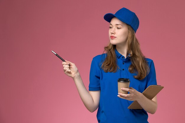 Weiblicher Kurier der Vorderansicht in der blauen Uniform, die braune Kaffeetasse mit Notizblock auf der hellrosa Hintergrunddienstuniform hält, die Firmenarbeit liefert