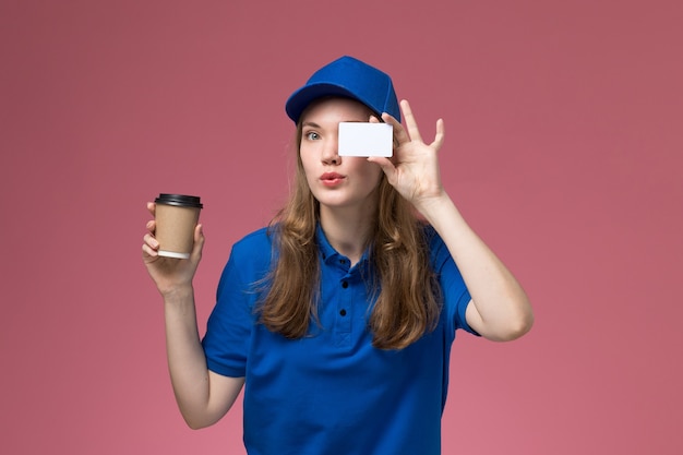 Weiblicher Kurier der Vorderansicht in der blauen Uniform, die braune Kaffeetasse mit weißer Karte auf hellrosa Schreibtischdienstjobuniformarbeitslieferfirma hält