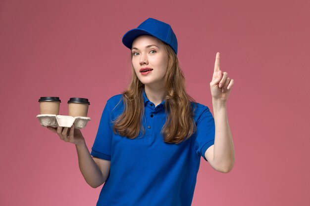 Weiblicher Kurier der Vorderansicht in der blauen Uniform, die braune Kaffeetassen auf der rosa Schreibtischdienstuniform-Lieferfirma hält