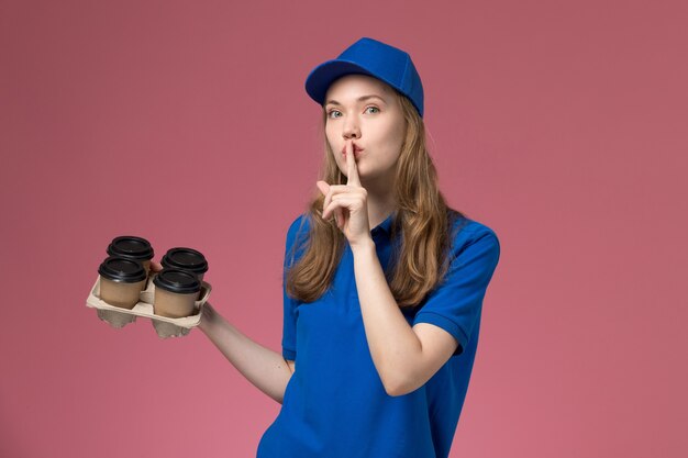 Weiblicher Kurier der Vorderansicht in der blauen Uniform, die braune Kaffeetassen der Lieferung hält, die Schweigenzeichen auf rosa Firmenservice der Schreibtischdienstuniform zeigen