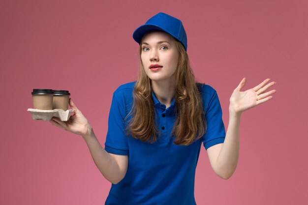 Weiblicher Kurier der Vorderansicht in der blauen Uniform, die braune Kaffeetassen mit verwirrtem Ausdruck auf der rosa Schreibtischdienstuniform hält, die Firmenjob liefert