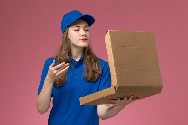 Weiblicher Kurier der Vorderansicht in der blauen Uniform, die Lebensmittel-Lieferbox hält, die es auf dem rosa Firmenarbeitsuniform-Firmenjob des Schreibtischarbeiters riecht