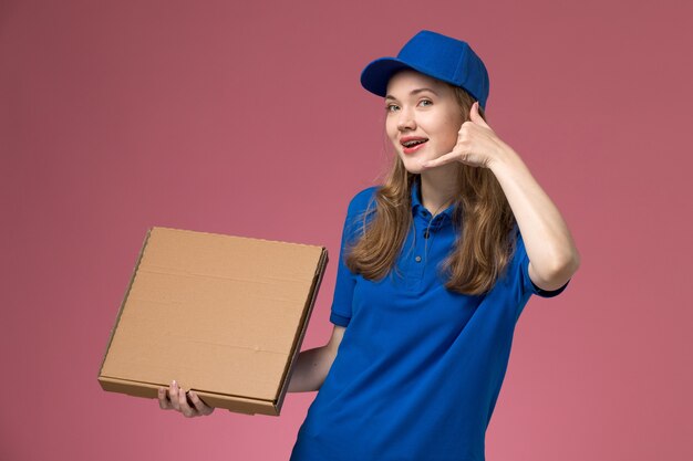 Weiblicher Kurier der Vorderansicht in der blauen Uniform, die Nahrungsmittelbox hält und auf rosa Hintergrundjobdienstuniformfirma lächelt