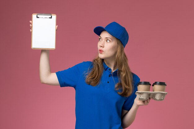 Weiblicher Kurier der Vorderansicht in der blauen Uniform, die Notizblock und Lieferkaffeetassen auf Lichtschreibtischdienstuniformfirma hält