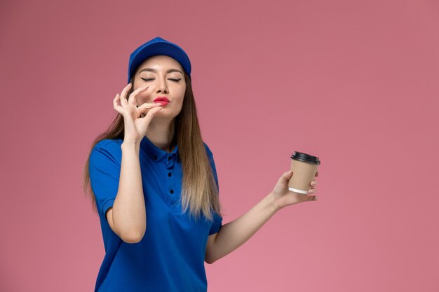 Weiblicher Kurier der Vorderansicht in der blauen Uniform und im Umhang, der die Kaffeetasse der Lieferung auf der rosa Wandjobmädchenarbeiterin hält