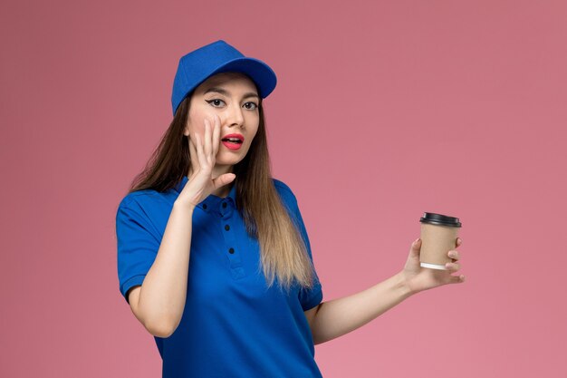 Weiblicher Kurier der Vorderansicht in der blauen Uniform und im Umhang, der die Kaffeetasse der Lieferung hält, die auf dem rosa Wandjobmädchen flüstert