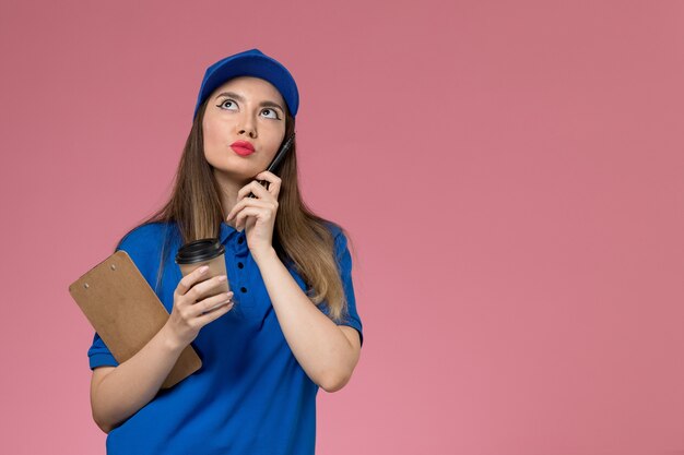 Weiblicher Kurier der Vorderansicht in der blauen Uniform und im Umhang, der die Kaffeetasse der Lieferung hält, die auf hellrosa Wand denkt
