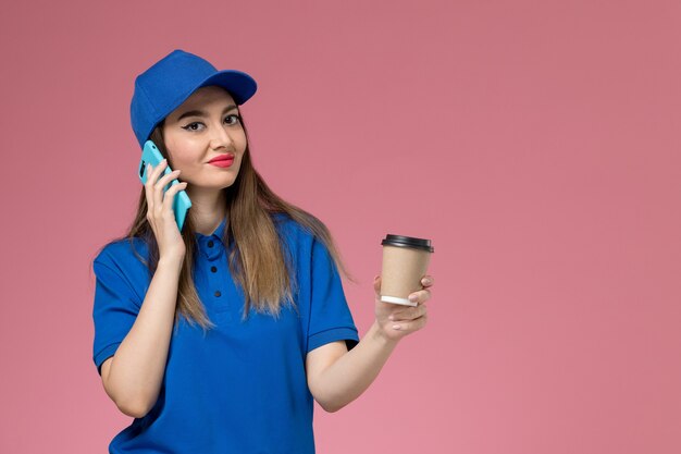 Weiblicher Kurier der Vorderansicht in der blauen Uniform und im Umhang, der die Kaffeetasse der Lieferung hält und am Telefon auf rosa Schreibtisch spricht