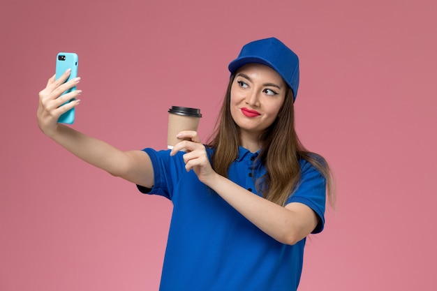 Weiblicher Kurier der Vorderansicht in der blauen Uniform und im Umhang, der die Kaffeetasse der Lieferung hält und Foto mit ihm auf der rosa Wand macht