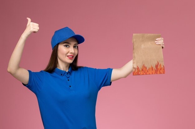 Weiblicher Kurier der Vorderansicht in der blauen Uniform und im Umhang, der Papiernahrungsmittelpaket hält, das auf rosa Wand lächelt