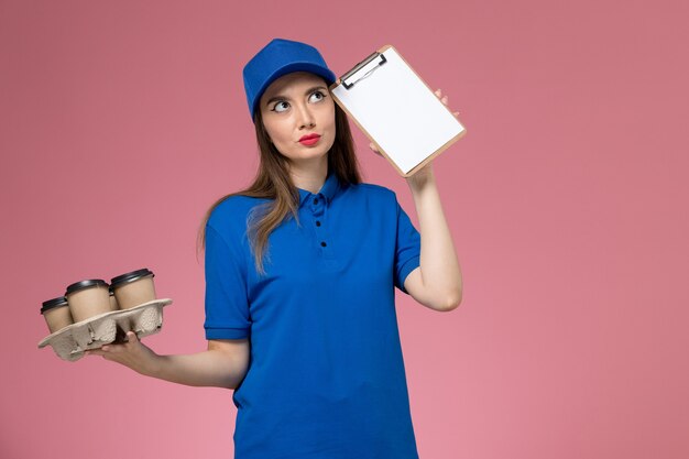 Weiblicher Kurier der Vorderansicht in der blauen Uniform und im Umhang, die Kaffeetassen mit Notizblock denken auf rosa Wandarbeiter halten